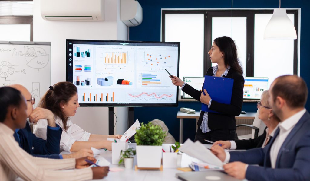 Young team leader in big corporation briefing coworkers pointing at graph meeting. Corporate staff discussing new business application with colleagues looking at screen